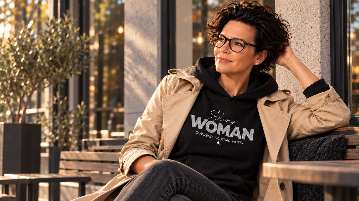 Frau mit kurzer dunkler Frisur und Brille trägt den schwarzen SHINY WOMAN Hoodie unter einem beigen Trenchcoat im warmen Abendlicht.  ECHT.STARK.UNZENSIERT. | alle-KAPUTT