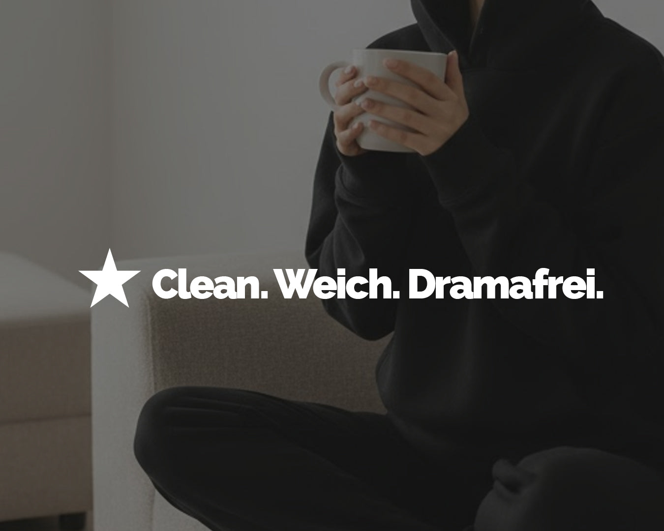 Frau im schwarzen Hoodie sitzt entspannt auf einem hellen Sofa und hält eine Tasse. Bildausschnitt ohne Gesicht.