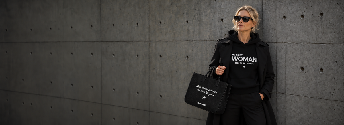 Frau mit Sonnenbrille im schwarzen ME FIRST WOMAN Hoodie mit weißem Print, schwarzem Mantel und Statement-Tasche von alle-KAPUTT vor einer Betonwand. ICH. KLAR. EIGEN.