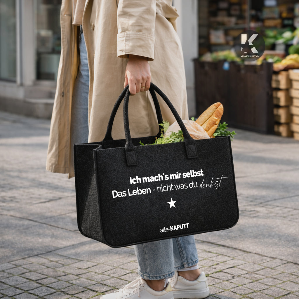 Schwarze Filztasche mit Statement „Ich mach’s mir selbst. Das Leben – nicht was du denkst.“ gefüllt mit Einkäufen auf dem Wochenmarkt, getragen von einer Frau, Marke alle-KAPUTT.