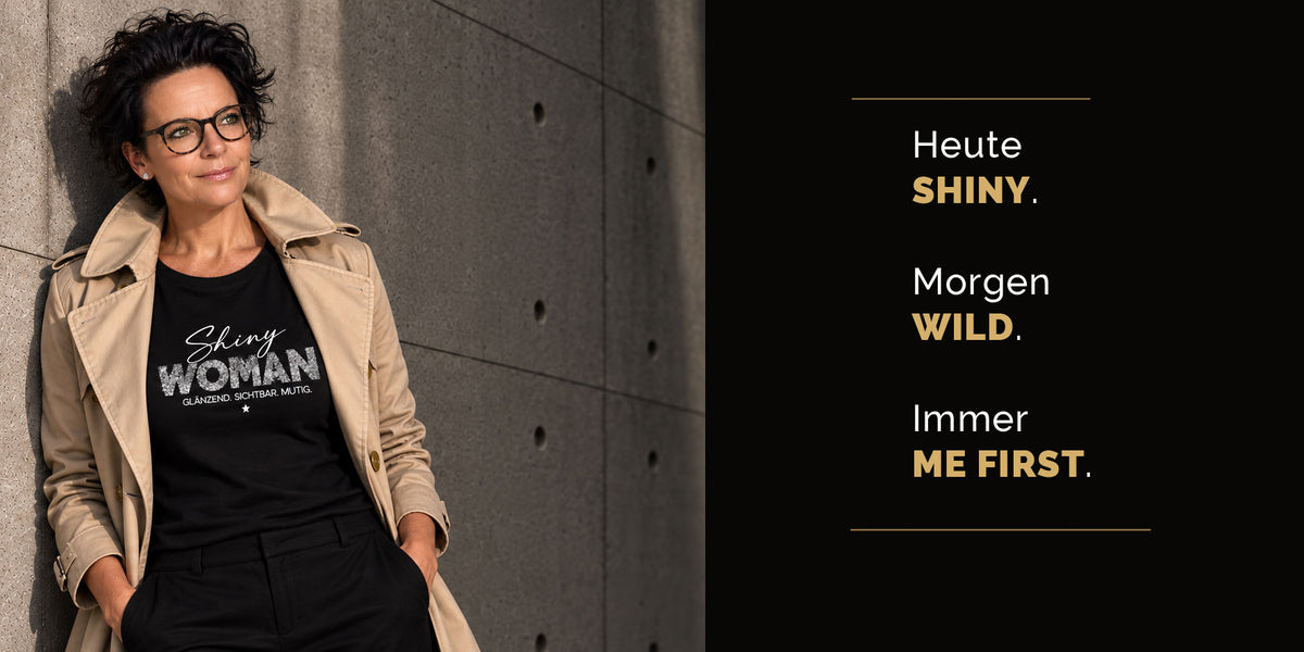 Frau mit kurzen dunklen Haaren im beigen Trenchcoat trägt schwarzes "Shiny Woman"-T-Shirt mit Glitzer-Print, lehnt an Betonwand – Hero-Bild für alle-KAPUTT Kollektionen Shiny, Wild, ME FIRST WOMAN