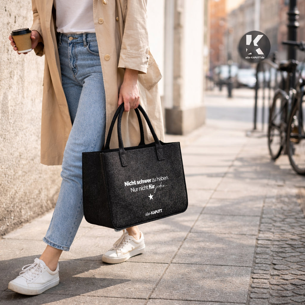 Schwarze Filztasche mit dem Statement „Nicht schwer zu haben. Nur nicht für jeden.“ getragen von einer Frau im Trenchcoat auf der Straße – alle-KAPUTT Lifestyle Bild