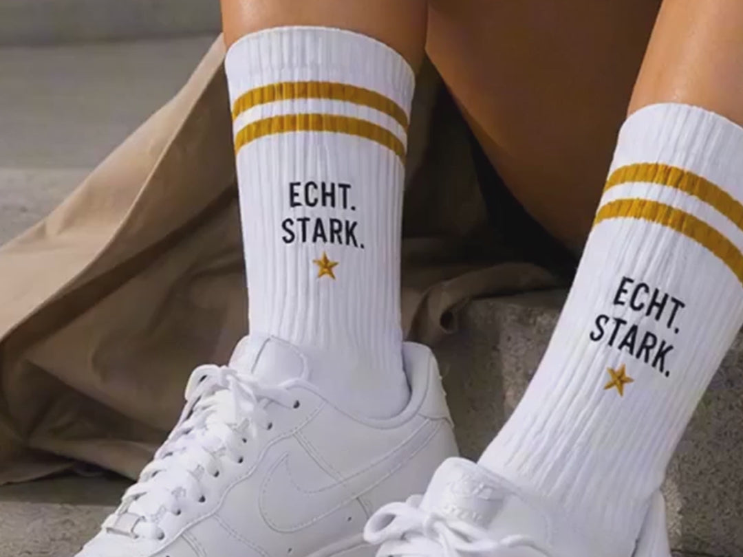 Weiße alle-KAPUTT Statement-Socken mit goldenen Streifen und dem Schriftzug „ECHT. STARK.“ in weißen Sneakern auf Betonstufen.