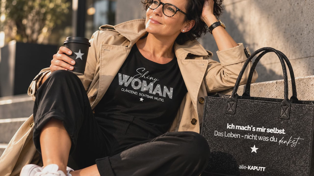 Selbstbewusste Frau im beigen Trenchcoat sitzt lässig auf Steinstufen und trägt ein schwarzes „Shiny Woman“-Shirt. Neben ihr steht eine schwarze alle-KAPUTT-Tasche mit Statement-Print in urbaner Lifestyle-Kulisse.